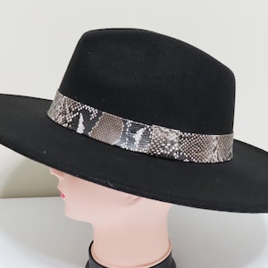 May include: A black wide-brimmed fedora hat featuring a structured crown and a flat brim. The base of the crown is encircled by a decorative band with a brown, grey, and white snakeskin pattern that contrasts against the solid black felt material.
