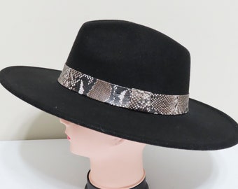 Puede incluir: Un sombrero fedora negro de ala ancha con una copa estructurada y ala plana. La base de la copa está rodeada por una banda decorativa con un patrón de piel de serpiente en tonos marrón, gris y blanco que contrasta con el fieltro negro sólido.