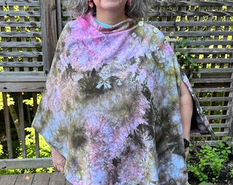 May include: A poncho featuring a tie-dye pattern in shades of pink, light blue, and dark olive green. The garment has a draped neckline and is secured along the shoulder with three white buttons. The fabric displays a mottled, organic design.