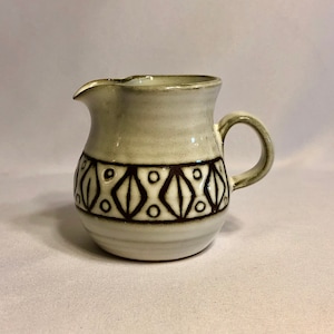 May include: A small, white ceramic creamer with a brown geometric pattern. The creamer has a rounded body and a handle.