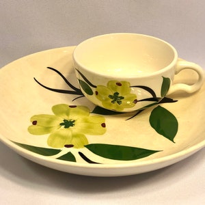 May include: A white ceramic teacup and saucer set with a floral design. The cup and saucer feature green leaves and yellow flowers with red centers.
