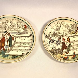 May include: Two vintage ceramic plates with colorful illustrations and musical notes. The first plate features a scene of people fishing with the text "LA MUETTE DE PORTICI" and musical notes. The second plate features a scene of people in military uniforms with the text "MIGNON" and musical notes.