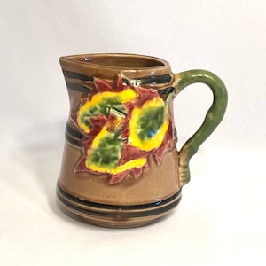 May include: A brown ceramic creamer with a green handle and a raised design of yellow and red leaves.