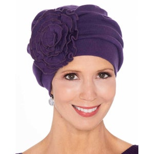 May include: Purple fabric hat with a large purple fabric flower on the side. The hat is soft and comfortable and is designed to be worn by people with hair loss.