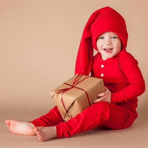May include: A child wearing a red long-sleeved onesie and a red elf hat holds a brown wrapped gift box with a red ribbon.