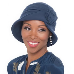 May include: A dark blue cloche hat featuring a soft, pleated crown and a decorative fabric rosette on the side. The hat has a downward-sloping brim that frames the face, crafted from a smooth, solid-colored material with a consistent matte finish.