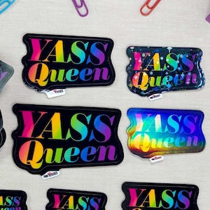 May include: A set of eight rainbow holographic stickers with the text "YASS QUEEN" in black. The stickers are arranged in a variety of positions on a light beige surface with colorful paper clips.