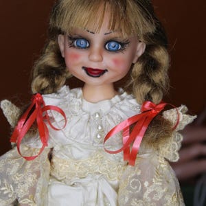May include: A vintage doll with a porcelain face, blue eyes, and red lips. The doll has blonde hair styled in pigtails with red ribbons. She wears a white dress with lace sleeves and a pearl-adorned neckline. The doll's cheeks are blushed.