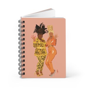 May include: A pink spiral bound notebook with an illustration of two women in colorful outfits. The woman on the left is wearing a yellow and black leopard print outfit with a large feathered hat. The woman on the right is wearing an orange jumpsuit with a blonde updo.