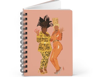 May include: A pink spiral bound notebook with an illustration of two women in colorful outfits. The woman on the left is wearing a yellow and black leopard print outfit with a large feathered hat. The woman on the right is wearing an orange jumpsuit with a blonde updo.