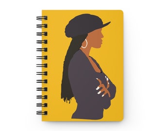 May include: A yellow notebook with a black spiral binding features a digital illustration of a woman with dark hair wearing a black hat and a dark gray jacket. The woman is looking to the right.