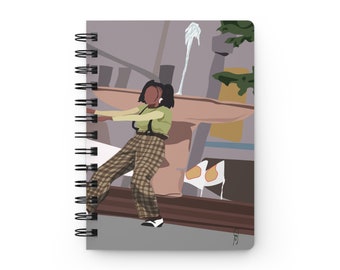 May include: A spiral bound notebook with a colorful illustration of a person wearing a plaid pants and a green shirt standing in front of a fountain. The background is a light gray with a brown and yellow structure.