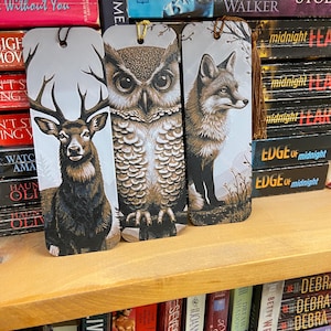 Può includere: Tre segnalibri decorativi con illustrazioni di animali selvatici. I segnalibri raffigurano un cervo, un gufo e una volpe, ognuno con opere d'arte dettagliate e un nappa. Sono esposti su una mensola di legno davanti a una collezione di libri.