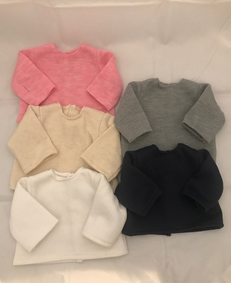 May include: Four long-sleeved shirts in different colors: pink, beige, gray, and navy blue. The shirts are folded and stacked on top of each other.