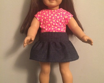 May include: A doll wearing a pink and white floral print shirt and a denim skirt. The doll has brown hair and is wearing black sandals.