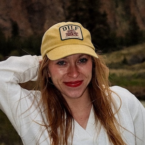 Puede incluir: Una mujer con una gorra de béisbol amarilla con el texto "DILF" y una sudadera con capucha blanca. Sonríe, con lápiz labial rojo, y está de pie al aire libre con un fondo escénico de montañas y un río.