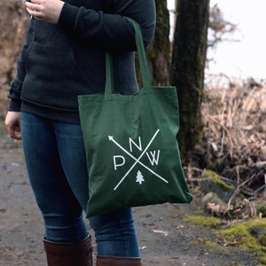 May include: A woman wearing a dark gray hooded sweatshirt with a white logo and blue jeans is holding a green tote bag with a white logo that says "NW" with a pine tree inside a circle.