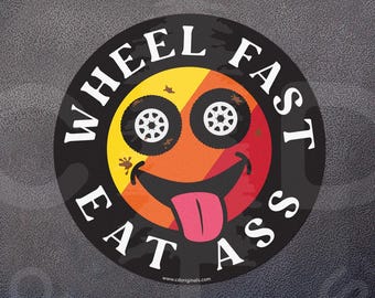 May include: A circular sticker featuring a yellow, orange, and red gradient background with a black border. The design includes two black tires as eyes, a wide black smiling mouth, and a pink tongue. The text WHEEL FAST EAT ASS is printed in white.