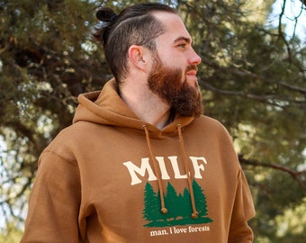 May include: A brown hoodie with a front pocket and a drawstring hood. The front of the hoodie features the word "MILF" in a cream-colored font above a green forest graphic, with the text "man, I love forests" below.