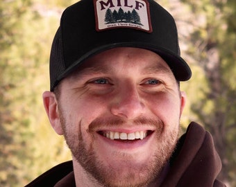 May include: Black trucker hat with a rectangular patch featuring the word "MILF" above a forest graphic and the text "man, I love forests". The hat is worn by a person wearing a brown hoodie.