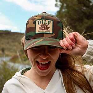Puede incluir: Una gorra de camionero de camuflaje con un parche verde que dice "DILF" y "dang, I love fungi!" en texto blanco.