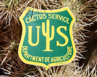 May include: A shield-shaped sticker with a green background and yellow border. The text reads CACTUS SERVICE and DEPARTMENT OF AGRICULTURE. In the center, the letters U and S flank a yellow cactus icon, mimicking a government agency emblem.
