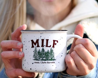 May include: White ceramic mug with black rim and speckled glaze. The mug has the text "MILF man, I love forests" in brown and green lettering with a graphic of five green trees.