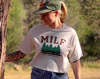 May include: A white t-shirt with a green forest graphic and the text "MILF man, I love forests". The person is wearing a green mesh baseball cap and blue denim shorts. They have tattoos on their legs.