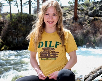 May include: A mustard yellow short-sleeve t-shirt featuring the green text DILF at the top, a brown graphic illustration of several mushrooms in the center, and the brown text dang, i love fungi printed underneath the mushroom illustration.