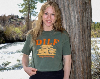 May include: Olive green crop top with the text "DILF" above a mushroom graphic and the phrase "dang, I love fungi!" in orange. The top is paired with black leggings. The person is leaning against a tree.