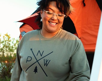 May include: A person wearing a green sweatshirt with a black logo that says "PNW" with a pine tree in the center. The person is standing in front of a tent and holding a can of soda.