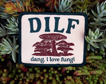 May include: A beige fabric patch with a green border. The patch features a dark green text that reads "DILF" and a brown illustration of mushrooms with the text "dang, i love fungi" below.