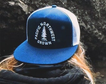 May include: A person wearing a blue and white trucker hat with the text "PACIFIC NORTHWEST GROWN" stands before a waterfall. The hat has a blue brim and a white mesh back. A black backpack is visible.