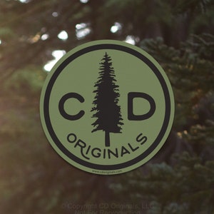 May include: A circular sticker with a green background and a black border. The sticker features the letters "C" and "D" flanking a black silhouette of a pine tree. The word "ORIGINALS" is printed below the tree in black. The website address is printed at the bottom.