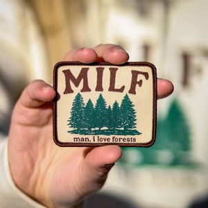 May include: A beige patch with brown stitching and the text "MILF" in brown letters. Below the text are five green trees and the text "man, i love forests".