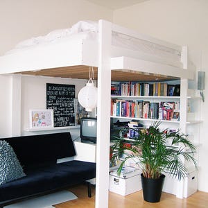 May include: A white loft bed with a white mattress and a white bookcase with books and a plant. The bed is in a small room with a black couch and a television.