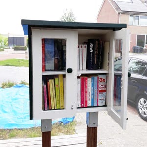 以下が含まれることがあります： さまざまな色とサイズの書籍で満たされた、濃い緑色の屋根とガラスのドアを備えた、小さな白い木製の無料図書館。 図書館は木製の柱に取り付けられており、屋外にあります。