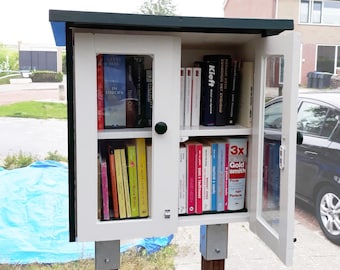 May include: A small, white wooden free library with a dark green roof and glass doors, filled with books of various colors and sizes. The library is mounted on wooden posts and is located outdoors.