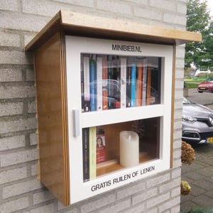 以下が含まれることがあります： ガラス張りの白い木製のブック交換ボックスで、「MINIBIEB.NL」と「GRATIS RUIEN OF LENEN」と書かれた看板が付いています。ボックスはレンガの壁に取り付けられており、本でいっぱいの棚があります。