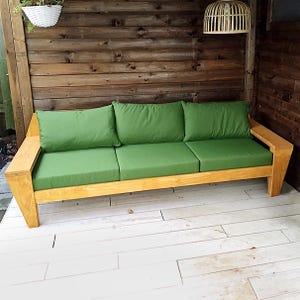 Op de afbeelding: Een houten buitenbank met een licht houten frame en drie groene kussens. De bank staat op een wit houten terras.