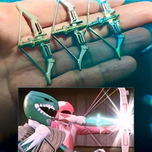 May include: Three miniature toy bows with silver, gold, and green accents are held in a hand. The bows have a futuristic design. A still from a show featuring two costumed characters aiming bows is in the lower portion of the image.
