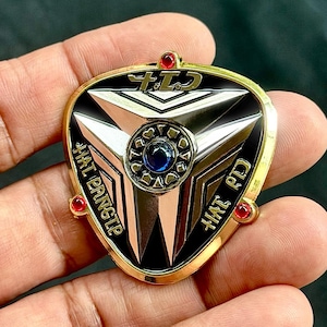 May include: A gold-toned metal badge with a black and silver geometric design. The badge has a central blue gem surrounded by a circular pattern. The text "HAT PRNSLP" and other Japanese characters are visible.