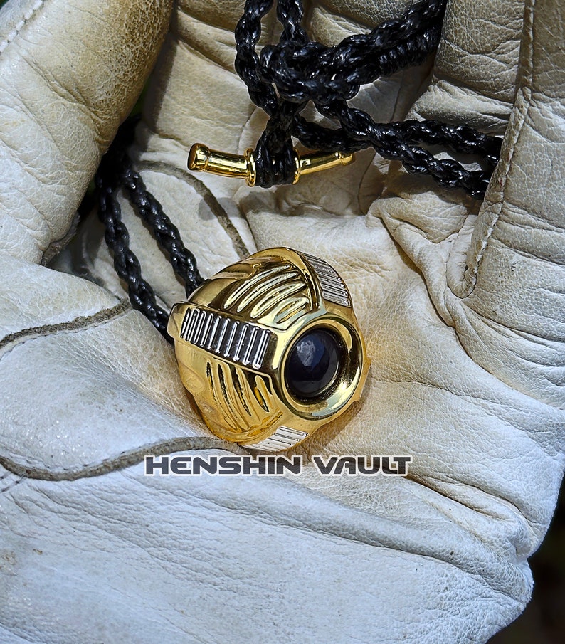 May include: A gold-coloured pendant with a black gem in the centre. The pendant is shaped like a sphere with intricate details and is attached to a black braided cord.