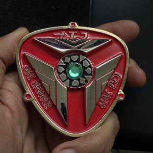 May include: A red and gold metal badge with silver accents. The badge has a triangular shape with the text "HAT BANGLE" and "HAT PD" in white. A green gem is in the center.