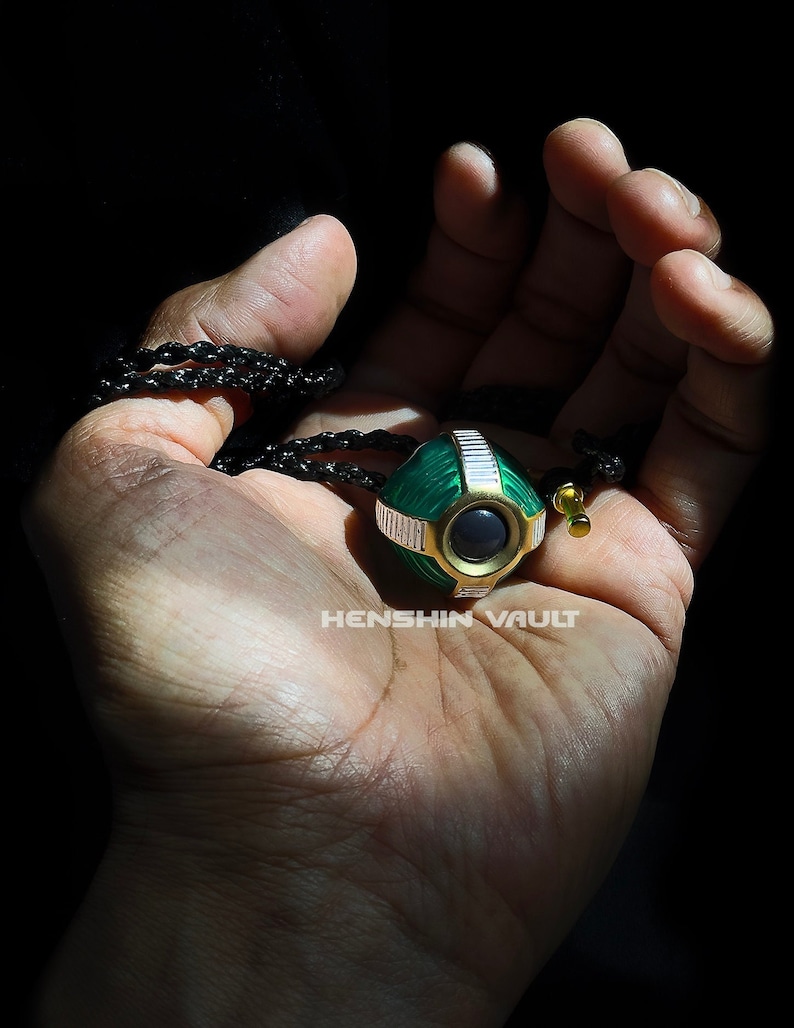 May include: A green and gold pendant with a black cord. The pendant is round with a black centre and white stripes. The text "HENSHIN VAULT" is visible on the pendant.