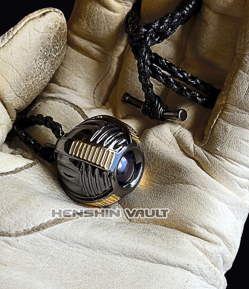 May include: A black and gold metal pendant shaped like a sphere with a lens on one side. The pendant is on a black cord and is being held in a white leather glove.