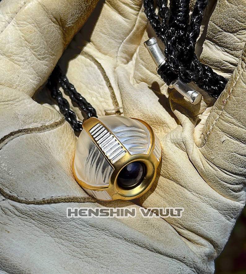 May include: A gold and silver pendant necklace with a round, futuristic-looking charm. The charm has a lens-like feature and a textured surface. The necklace is on a black cord.