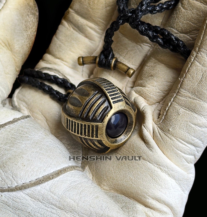 May include: A gold-toned metal pendant with a geometric design and a dark blue gemstone. The pendant is hanging from a black braided cord.
