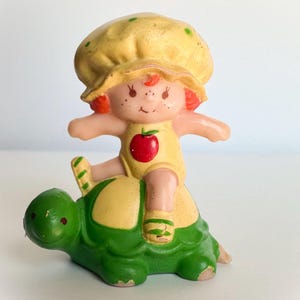 May include: A vintage-style plastic figurine featuring a small character with red hair wearing a yellow bonnet and a yellow outfit with a red apple graphic, riding on the back of a green turtle with a yellow shell pattern and painted details.