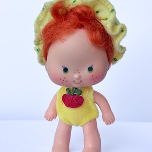 May include: A small plastic doll with bright orange hair, green eyes, and freckles on the cheeks. The doll wears a yellow ruffled bonnet with green dots and a yellow one-piece outfit featuring a red embroidered strawberry on the chest.